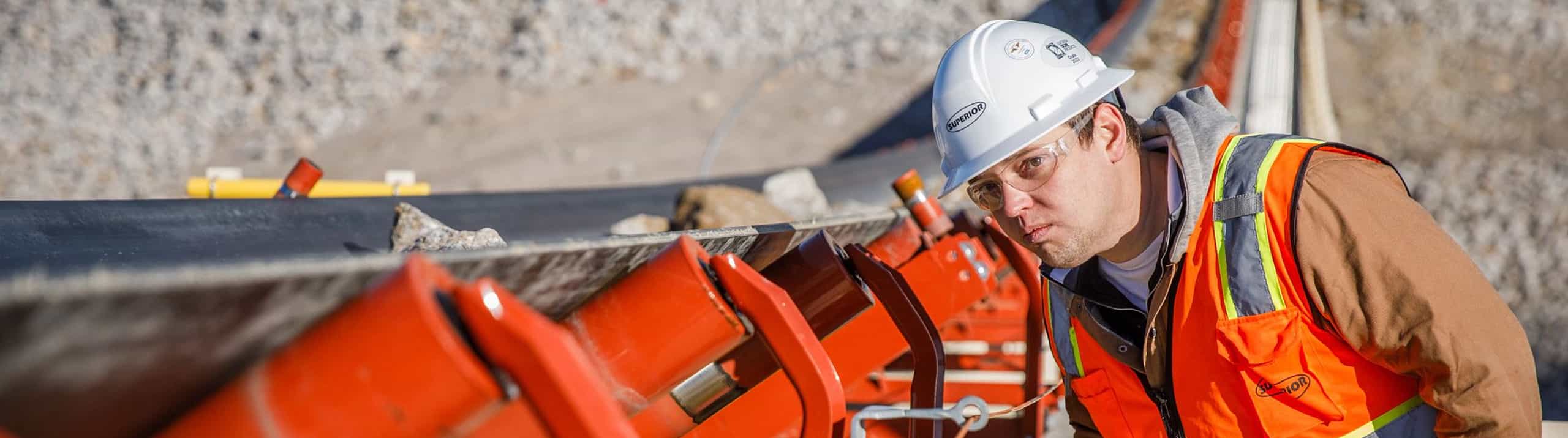 Operator with hard hat inspecting belt tracking and idlers on surge reclaim conveyor belt at quarry site.